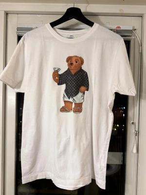 Vit t-shirt med björntryck - Vit t-shirt med ett stort tryck av en björn i skjorta och shorts på framsidan. Klassisk rund hals och korta ärmar. Tillverkad i mjuk bomull. 