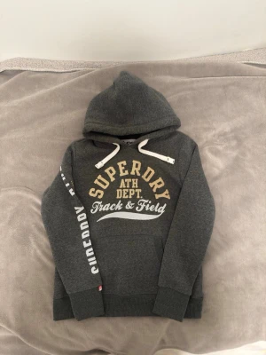 mörkgrön Superdry hoodie med tryck - Mörkgrå hoodie från Superdry med stor huva och vita dragsnören. Framsidan har gul och vit text: 'Superdry ATH DEPT Track & Field'. På ena ärmen står det 'Superdry ATHL.' i vitt och gult. Hoodien har magficka och ribbade muddar. Perfekt för en sportig och avslappnad stil. PRIS KAN DISKUTERARS