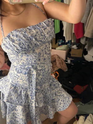 Blåvit blommig kortklänning med volang - Supersöt kortklänning med blått blommönster på vit botten. Klänningen har smala axelband, volangdetaljer och knyt i midjan. Perfekt för varma dagar och har en feminin, flowy passform.