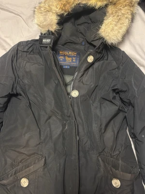 Svart parkas från Woolrich med päls - Svart parkas från Woolrich med huva och fuskpälsdetalj. Jackan har dragkedja och stora knappar framtill samt praktiska fickor. Perfekt för kalla dagar med sin långa passform och klassiska design.