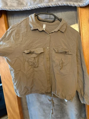 Beige croppad skjorta från FB Sister - Säljer en beige croppad skjorta från FB Sister med långa ärmar, två stora bröstfickor med lock och knytning i nederkanten. Skjortan har klassisk krage och knappar framtill. Perfekt för en avslappnad och trendig look.