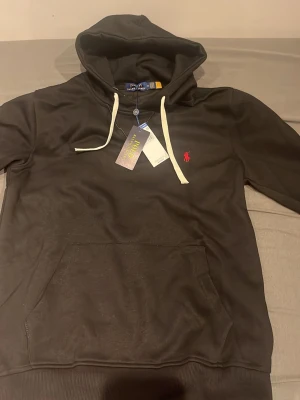 Svart hoodie från Polo Ralph Lauren - Svart hoodie från Polo Ralph Lauren med vit dragsko i huvan och klassisk röd logga broderad på bröstet. Hoodien har en stor magficka och ribbade muddar. Perfekt för en chill och stilren look.