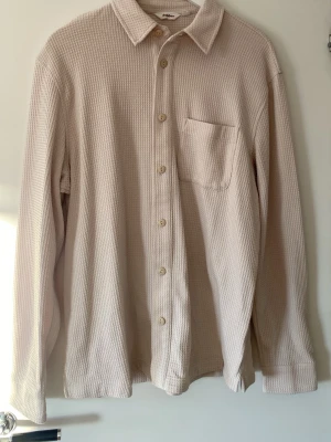 Beige skjorta i bomull med struktur från dobber  - Säljer en beige skjorta med lång ärm och knappar framtill i storlek M. Skjortan har en strukturerad, ribbad yta och en bröstficka. 