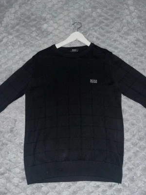 Svart tröja från Hugo Boss - Snygg svart tröja från Hugo Boss med diskret rutmönster och rund halsringning. Tröjan har en broderad logga på bröstet och ribbade muddar vid ärmslut och nederkant. 