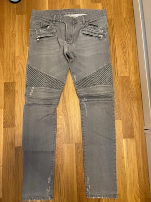 Gråa bikerjeans från Balmain - Säljer ett par grå bikerjeans från Balmain med ribbade detaljer över knäna och dragkedjor vid fickorna. Jeansen har en slim passform och är tillverkade i mjukt denimtyg. Perfekta för dig som vill ha en edgy och modern stil.