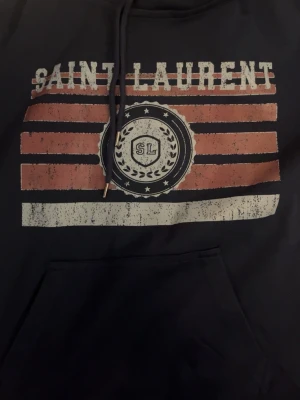 Mörkblå Saint Laurent hoodie - Mörkblå hoodie från Saint Laurent med tryck framtill i rött, vitt och svart. Stor magficka och justerbar huva med snören. Trycket har en vintage-look med texten 'Saint Laurent' och SL-emblem. Perfekt för en avslappnad och cool stil.