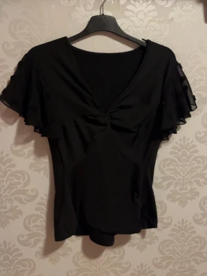 Svart blus/bluzka - Svart blus med v-ringning och volangärmar i transparent tyg med små dekorativa detaljer. Blusen har en lätt rynkning vid bysten och är figurnära i modellen. Perfekt att styla med jeans eller kjol.