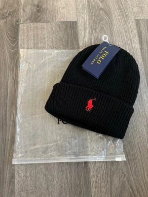 Svart mössa från Polo Ralph Lauren - Svart ribbstickad mössa från Polo Ralph Lauren med klassisk uppvikt kant och röd broderad logga framtill. Tillverkad i mjukt material som håller dig varm och snygg under kalla dagar. Perfekt accessoar för en stilren look.