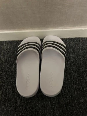 Vita Adidas Tofflor  med svarta ränder - Klassiska vita sandaler från Adidas med tre svarta ränder över foten. Tillverkade i syntetmaterial och har en platt sula samt öppen tå. Perfekta för sommaren och enkel att bara glida i.