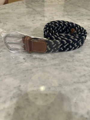 Flätat marinblått bälte med läderdetaljer - Snyggt flätat bälte i marinblått, grått och vitt med bruna läderdetaljer vid spännet. Spännet är i metall och har en klassisk form. Perfekt accessoar för att lyfta din outfit med en stilren touch.
