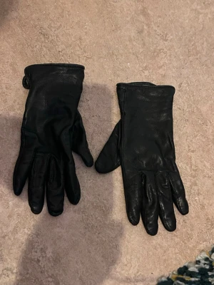Svarta skinnhandskar med ullfoder - Stilrena svarta handskar i äkta skinn med mjukt beige ullfoder på insidan. Klassisk design med knappdetalj vid handleden och storlek 7.0. Perfekta för kalla dagar när du vill hålla händerna varma och samtidigt se snygg ut.