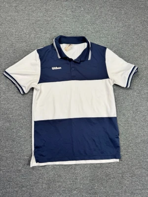 Wilson pikétröja blåvit - Snygg pikétröja från Wilson i blått och vitt med klassisk krage och knappar. Tröjan har korta ärmar med randiga muddar och är perfekt för sportiga aktiviteter. Materialet känns lätt och andas bra.
