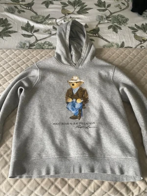 Grå hoodie Polo Bear Ralph Lauren - Grå hoodie från Ralph Lauren med Polo Bear-tryck framtill. Björnen har cowboy-outfit och texten 'Polo Bear by Ralph Lauren' under. Klassisk huva och ribbade muddar. Perfekt för dig som gillar streetstyle och ikoniska prints.