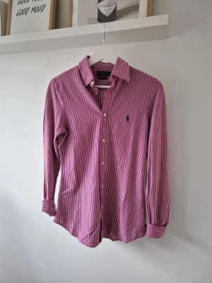 Rosa randig skjorta från Polo Ralph Lauren - Säljer en klassisk rosa skjorta från Polo Ralph Lauren med vita ränder och blå broderad logga på bröstet. Skjortan har lång ärm, knappar framtill och en tidlös krage. Perfekt för dig som gillar stilrena och snygga skjortor. 