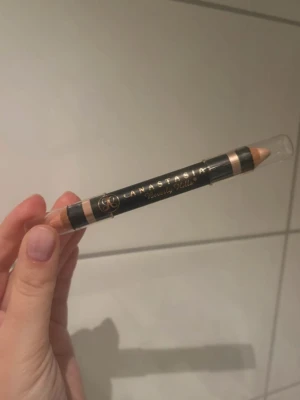 Anastasia Beverly Hills Duo Ögonbrynspenna - Dubbeländad ögonbrynspenna från Anastasia Beverly Hills med svart och guld text på hylsan. Ena sidan är ljusbeige och den andra är en något mörkare beige ton. Perfekt för att definiera och highlighta brynen. Pennan är i trä med plastlock.