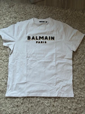 Vit Balmain Paris t-shirt - Snygg vit t-shirt från Balmain Paris med klassisk rund hals och svart textlogga framtill. T-shirten har korta ärmar och är gjord i mjuk bomull för en skön känsla. Perfekt för dig som vill ha en clean och trendig look.