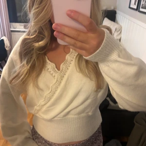 Vit omlottopp med spetsdetaljer - Säljer en söt vit omlottopp med långa ärmar och fina spetsdetaljer längs den djupa v-ringningen. Tröjan har bred ribbad midja som ger en snygg siluett och är perfekt för dig som gillar romantisk stil.