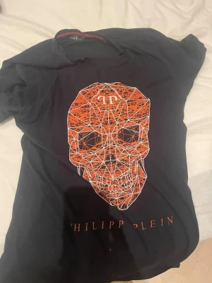 Svart  t-shirt med döskalle - Säljer en svart t-shirt från Philipp Plein med en stor grafisk döskalle i orange och vitt på bröstet. T-shirten har korta ärmar och rund hals. Materialet känns som bomull och trycket ger en cool och edgy vibe.