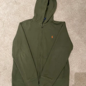 Olivgrön hoodie från Polo Ralph Lauren - Olivgrön hoodie från Polo Ralph Lauren med dragkedja och huva. Klassisk orange logga broderad på bröstet. Tillverkad i mjuk bomull, perfekt för chill dagar. Snygg och enkel att matcha med jeans eller joggers.