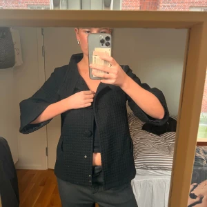 Svart kappa med trekvartsärm - Stilren svart kappa med markerad krage och stora knappar framtill. Jackan har ett diskret rutmönster i tyget och trekvartsärmar som ger en cool och modern vibe. Perfekt för dig som gillar klassiska plagg med en twist.