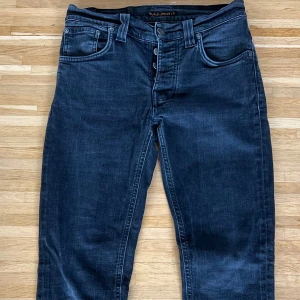 Nudie Jeans Grim Tim svarta jeans 30/32 - Svarta Grim Tim jeans från Nudie Jeans i storlek 30/32. Tillverkade i 97% ekologisk bomull och 3% elastan för en skön stretch. Klassisk femficksmodell med knappgylf och snygg tvättad finish. Perfekta för dig som gillar stilrena och bekväma jeans.