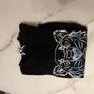 Svart Kenzo t-shirt med tigerbrodyr - Svart t-shirt från Kenzo med ikonisk broderad tiger i vitt, blått, orange och grönt framtill. Klassisk passform, rund hals och korta ärmar. Materialet är mjuk bomull och trycket har coola färgdetaljer som sticker ut. Perfekt statement-plagg för dig som gillar streetstyle. Passar även M