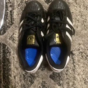 Adidas Superstar svart/vit strl 38  - Klassiska Adidas Superstar sneakers i svart skinn med vita ränder och guldiga detaljer på plösen. Skorna har snörning, rund tå och den ikoniska gummitå-designen. Perfekta för dig som gillar streetstyle och tidlös design. Kan dra ner priset ingen box