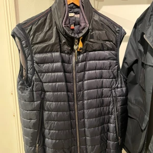 Svart dunväst från Parajumpers - Snygg svart dunväst från Parajumpers med quiltad design och hög krage. Västen har dragkedja framtill, två sidofickor med dragkedja och en ikonisk patch på ryggen. Perfekt för lager-på-lager och stilren look.