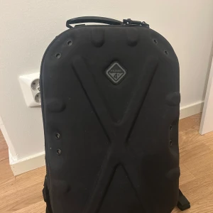 Svart ryggsäck från Hazard 4 - Säljer en svart ryggsäck från Hazard 4 med hård, formpressad framsida och coola detaljer. Ryggsäcken har flera fästen, justerbara remmar och vadderad baksida för extra komfort. Perfekt för dig som gillar funktionell och stilren design.