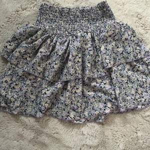 Blommig volangkjol från Gina Tricot - Supersöt kort volangkjol från Gina Tricot i blommigt mönster med lila, svart och vita toner. Kjolen har smock i midjan och dubbla volanger som ger en riktigt fin och flowig look. Perfekt för dig som gillar romantisk och trendig stil.