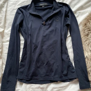 Marinblå långärmad tröja från Equipage - Marinblå långärmad tröja från Equipage med halv dragkedja framtill och hög krage. Perfekt för ridning eller träning, tillverkad i mjukt och stretchigt material som andas. Snygg och stilren design med en diskret logga på bröstet.