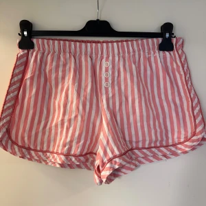 Randiga shorts från Bondelid strl: M - Säljer ett par rosa och vita randiga shorts från Bondelid. De har elastisk midja, tre dekorativa knappar framtill och broderad logga. Shortsen är i mjuk bomull och har en loose passform, perfekta för varma dagar eller till att sova med dom. Finns litet hål invändigt vid resåret. Förekommer noppor annars o fint skick.