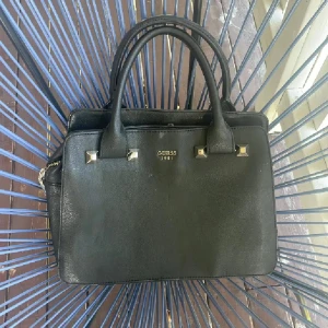 Svart handväska från Guess - Stilren svart handväska från Guess med guldfärgade detaljer och logotyp framtill. Väskan har två rejäla handtag, flera fack och dragkedja upptill. Tillverkad i skinnimitation med en klassisk, fyrkantig form som passar till många olika looks. Se defekterna på bilderna😁Endast swish med spårbar 59 eller mötas❤️
