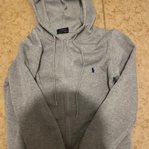 Grå hoodie från Polo Ralph Lauren - Snygg grå hoodie från Polo Ralph Lauren med dragkedja framtill och klassisk blå logga på bröstet. Tröjan har huva med snörning och långa ärmar. Perfekt för chill dagar eller när du vill ha en clean och enkel stil. Passad även m