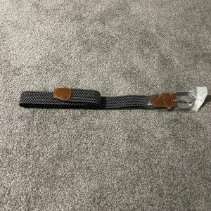 Svart och vit flätat bälte med skinn - Snyggt svart och vitt flätat bälte med bruna skinn-detaljer vid spännet och änden. Bältet har ett klassiskt mönster och ett silvrigt metallspänne. Passar perfekt till jeans eller chinos för en stilren look.