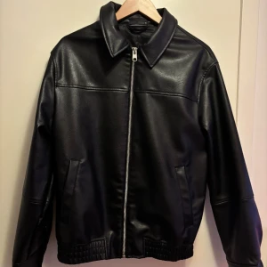 Svart skinnjacka från Zara, M - Snygg svart skinnjacka från Zara i storlek M. Jackan har klassisk krage, raka linjer och dragkedja framtill. Två sidofickor och ribbad nederkant ger en cool vibe. Materialet är syntetiskt skinn med lätt textur. Perfekt för dig som gillar stilrena och tidlösa plagg. Ny utan prislapp, använd enbart 2 gånger max.