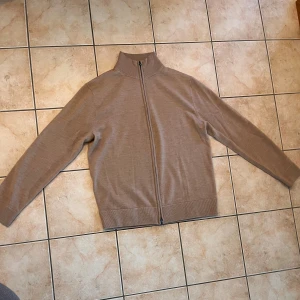 Beige merinoull kofta från These Glory Days - Snygg beige kofta i merinoull från These Glory Days. Tröjan har hel dragkedja framtill, ribbade muddar och hög krage. Perfekt för dig som gillar stilrena och klassiska plagg med skön passform. Storlek L och tillverkad i Superior Merino. NÄSTAN HELT NY SITTER SOM M 