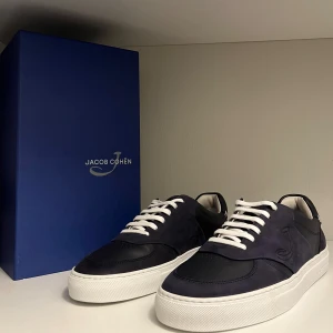 Jacob Cohën Sneakers  - Jacob Cohën Dreamer i en stilren mörkblå mocka och skinn kombination, med vit sula och en diskret J-logga ger dig exklusivitet, stil och komfort i ett. Perfekt för dig som gillar exklusiva och cleana sneakers. Går ej att få tag på längre dessa är helt nya box och dust bag ingår.    Nypris 7400kr Vårt pris: 6300kr