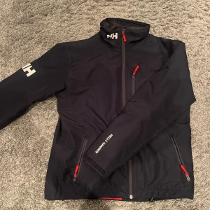 Svart vindjacka från Helly Hansen - Svart vindjacka från Helly Hansen med röda dragkedjor och vita logotryck på ärm och krage. Jackan är i Perfekt skick 