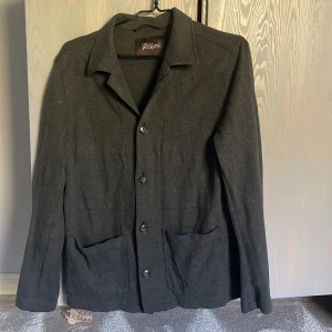 Mörkgrå overshirt från Oscar Jacobson - Snygg mörkgrå overshirt från Oscar Jacobson med klassisk krage, stora framfickor och knäppning med fyra knappar. Jackan har lång ärm och är tillverkad i mjuk ullblandning som ger en chill och stilren vibe. Perfekt att slänga över en hoodie eller t-shirt.