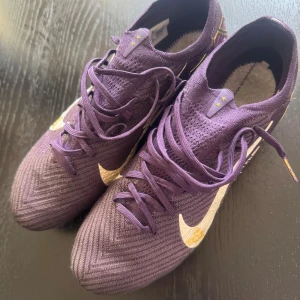 Nike Air Zoom fotbollsskor lila/guld - Säljer ett par Nike Air Zoom fotbollsskor i mörklila med guldiga detaljer och vit swoosh. Skorna har snörning, stickad ovandel och mönstrad sula med dobbar för gräs. Snyggt grafiskt mönster och coola detaljer som 'BALLER' och '93' på sidan. Perfekt för dig som vill sticka ut på planen. Dom är använda 3 gånger 