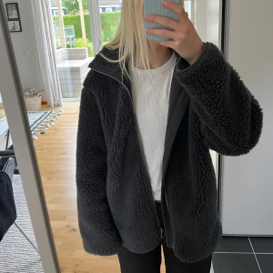 Mörkgrå teddyjacka från Weekday XS - Mysig svart teddyjacka från Weekday i storlek XS. Jackan har en hög krage, dragkedja framtill och är tillverkad i fluffigt syntetmaterial. Perfekt för kalla dagar.