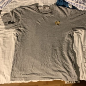 Grå t-shirt Comme des Garçons Play - Grå t-shirt från Comme des Garçons Play med klassisk rund hals och korta ärmar. På bröstet sitter det ikoniska hjärtat i guld med ögon, broderat. T-shirten är i mjuk bomull och har en clean, stilren look som passar perfekt till jeans eller shorts.