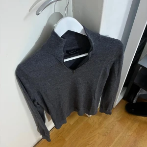 Grå merinoulltröja med half zip från Riley - Snygg mörkgrå långärmad tröja från Riley i mjuk merinoull. Tröjan har en stilren half zip-detalj vid halsen och en normal passform. Perfekt för dig som gillar minimalistisk och clean stil.