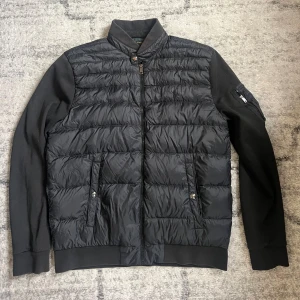 Svart pufferjacka från Ralph Lauren - Svart pufferjacka från Ralph Lauren med quiltad front, släta ärmar och ståkrage. Jackan har dragkedja, två fickor med knappstängning och en ärmficka med dragkedja. Fodret har ett mörkgrönt rutigt mönster. Perfekt för kyliga dagar.