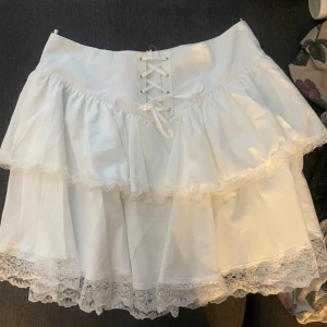 Vit volangkjol med snörning och spets - Supersöt vit kortkjol med två lager volanger och spetskant. Kjolen har snörning framtill i midjan för extra detalj och en feminin look. Perfekt för dig som gillar romantisk och lekfull stil. 