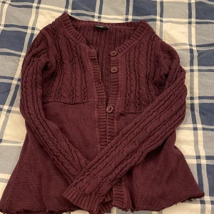 Vinröd kofta från Isolde, kabelstickad - Mysig vinröd kofta från Isolde med kabelstickat mönster och snygga knappar framtill. Tröjan har långärmade ärmar och ribbade detaljer vid halsringning och muddar. Perfekt för lager-på-lager och chill höstkvällar. OBS: En knapp saknas! 💕 priset kan såklart diskuteras! 