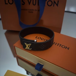 Louis Vuitton Monogram Armband - Stilrent armband från Louis Vuitton i svart skinn med klassiskt monogrammönster och guldfärgad LV-logga framtill. Armbandet har en enkel och snygg design som ger en lyxig känsla. Perfekt accessoar för dig som gillar exklusiva detaljer. Kvitto bevis finns! Box tillbehör medföljer!