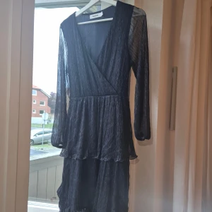 Glittrig omlottklänning från Kappahl - Supersnygg långklänning från Kappahl i mörkblått glittrigt material. Klänningen har omlottmodell med v-ringning, långa transparenta ärmar och volangdetaljer i kjolen. Perfekt för dig som vill sticka ut med lite extra shimmer och cool vibe.