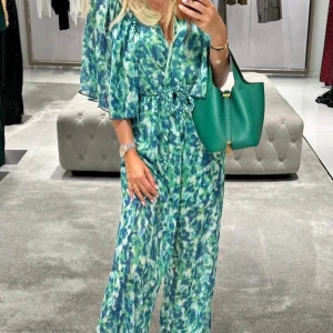 Blommig långklänning med knyt i midjan - Säljer en flowy långklänning med grönt och blått blommigt mönster. Klänningen har v-ringning, knyt i midjan och luftiga, korta ärmar. Materialet är lätt och känns som polyester, perfekt för varma dagar. Den har en avslappnad passform och ger en fräsch vibe.